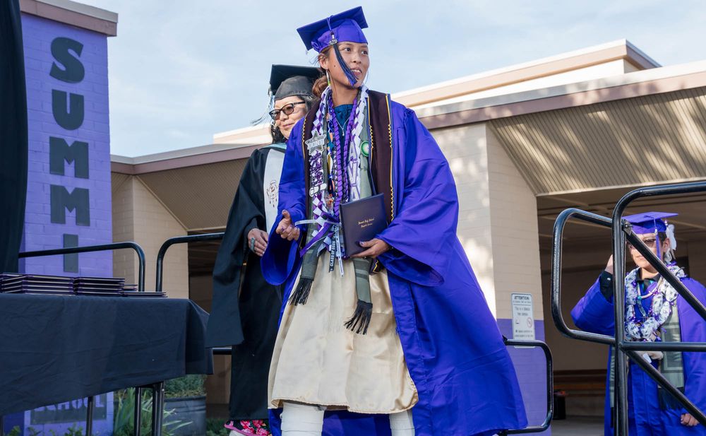 A graduate in bright blue regalia celebrates on stage during an outdoor commencement ceremony.