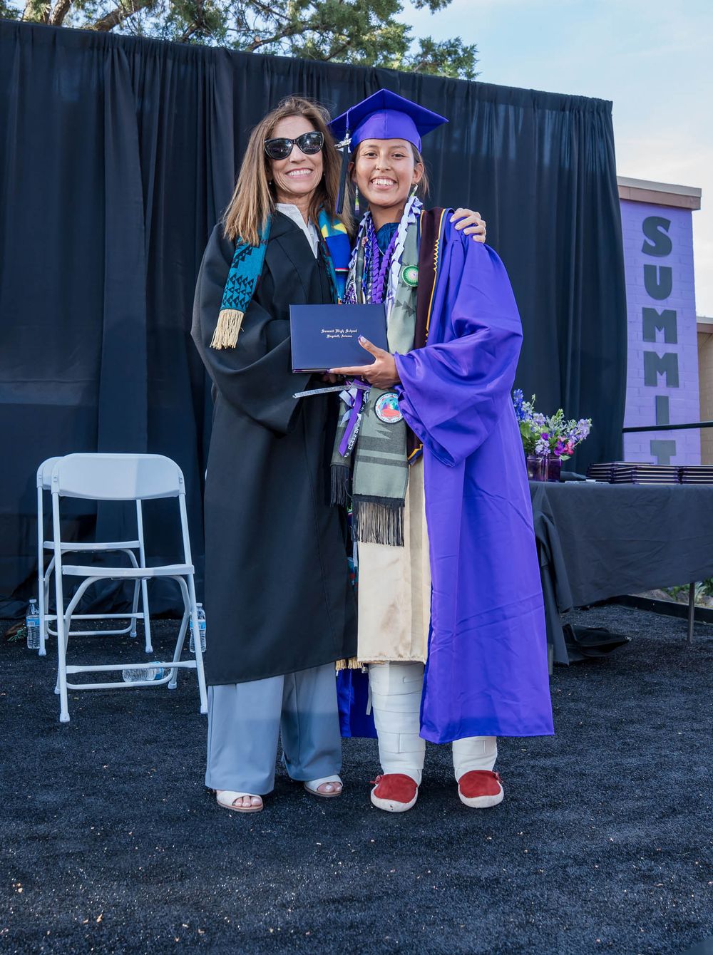 Two people in graduation attire pose together at a ceremony, one in black robes and one in bright purple regalia.