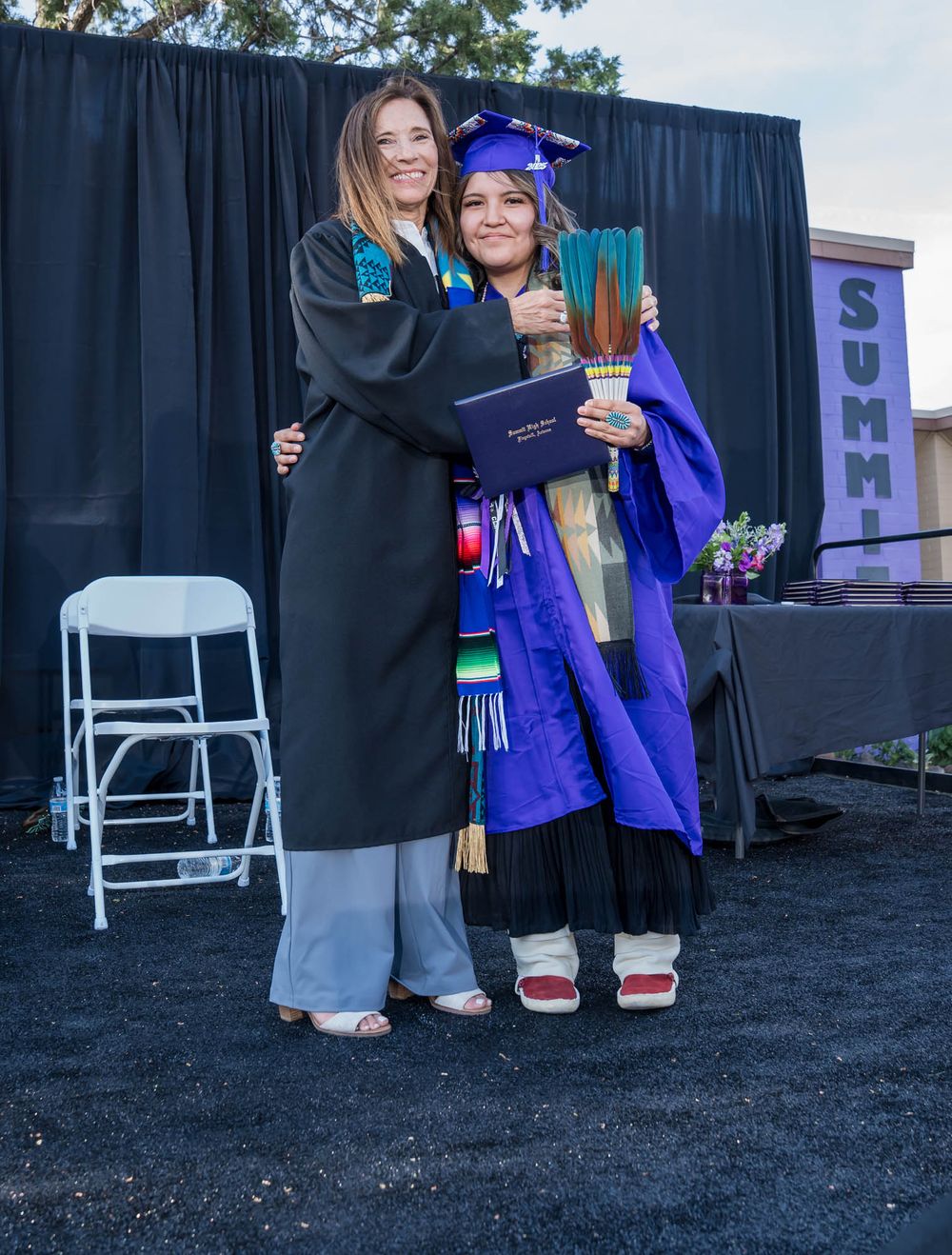 Two people in graduation regalia standing together at a ceremony, one in blue and one in black gowns, with a dark curtain backdrop.