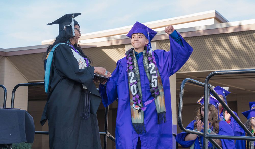 Two graduates in bright blue and black robes celebrate during an outdoor commencement ceremony on stage.