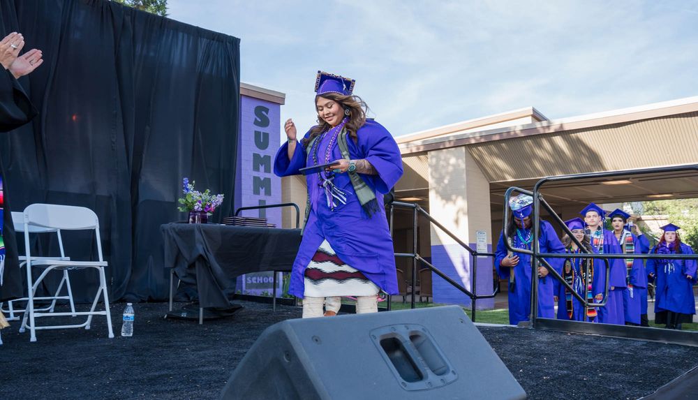 Graduate wearing royal blue regalia celebrates on outdoor stage during graduation ceremony.