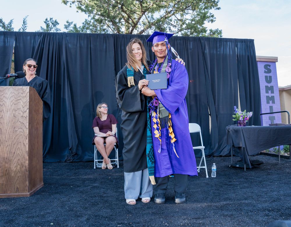 Graduation ceremony on outdoor stage with black backdrop and graduates in traditional academic regalia.