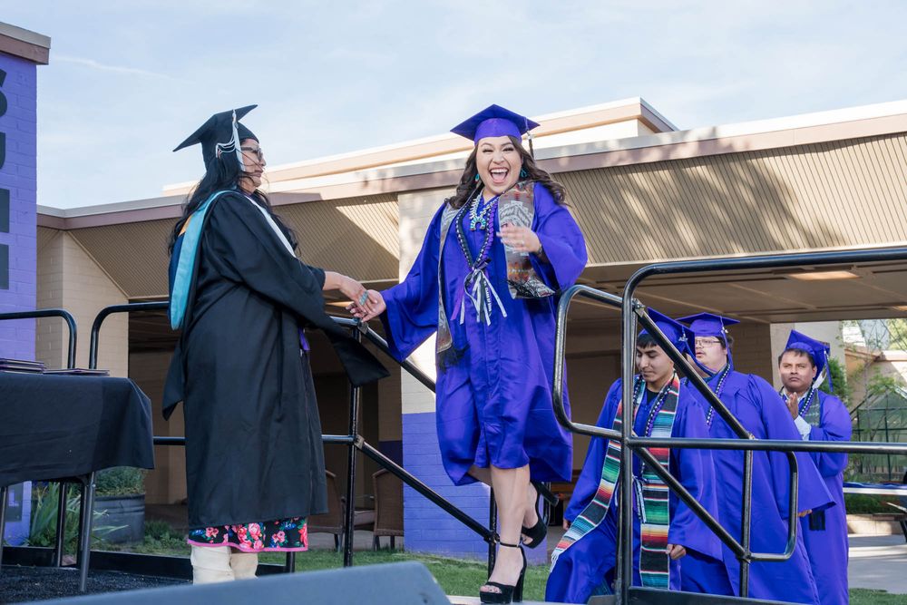 Graduate in blue cap and gown receives diploma on stage at outdoor graduation ceremony.