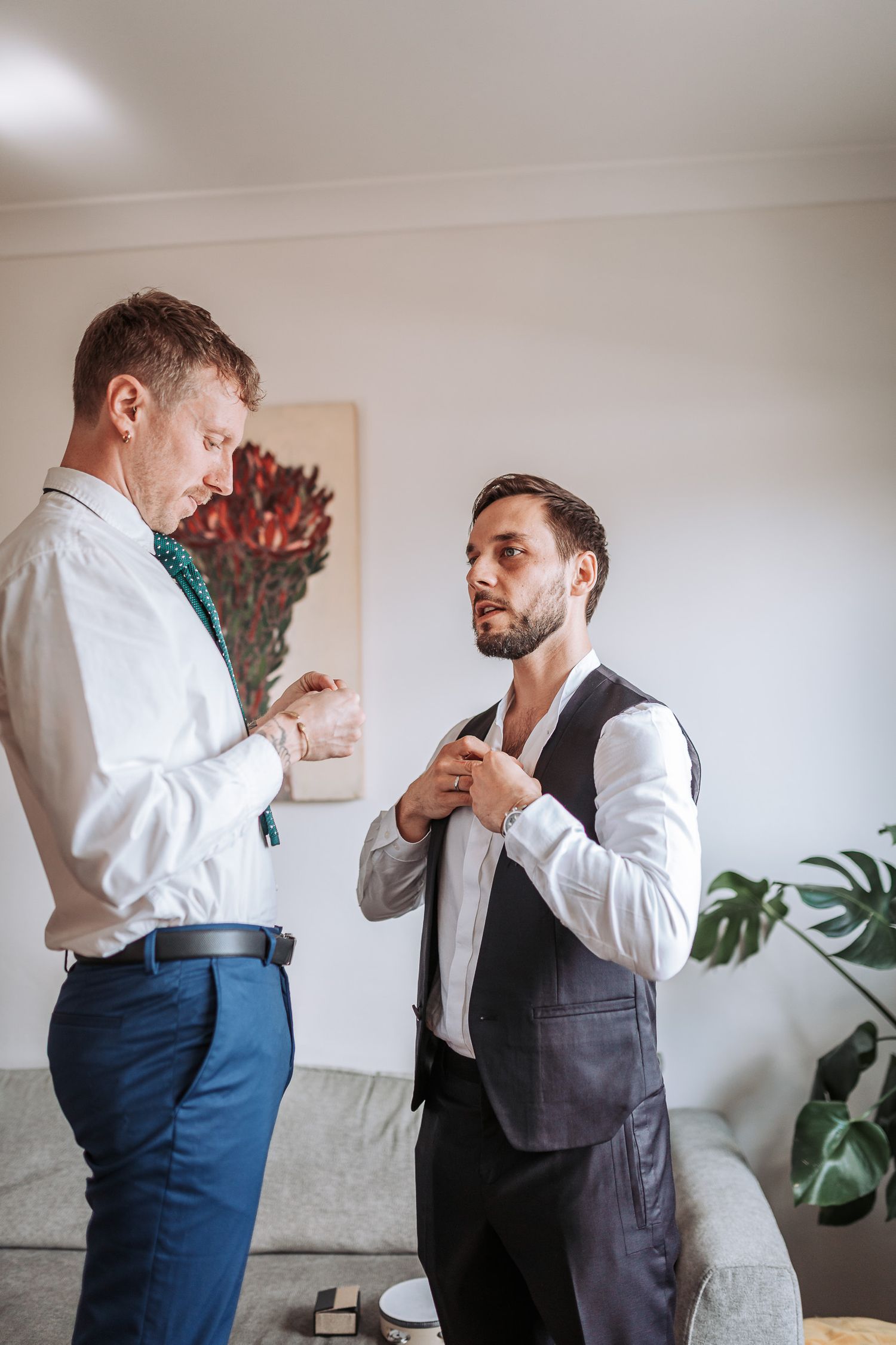 Two men in formal attire adjusting their ties and getting ready in a room with houseplants and wall decor.