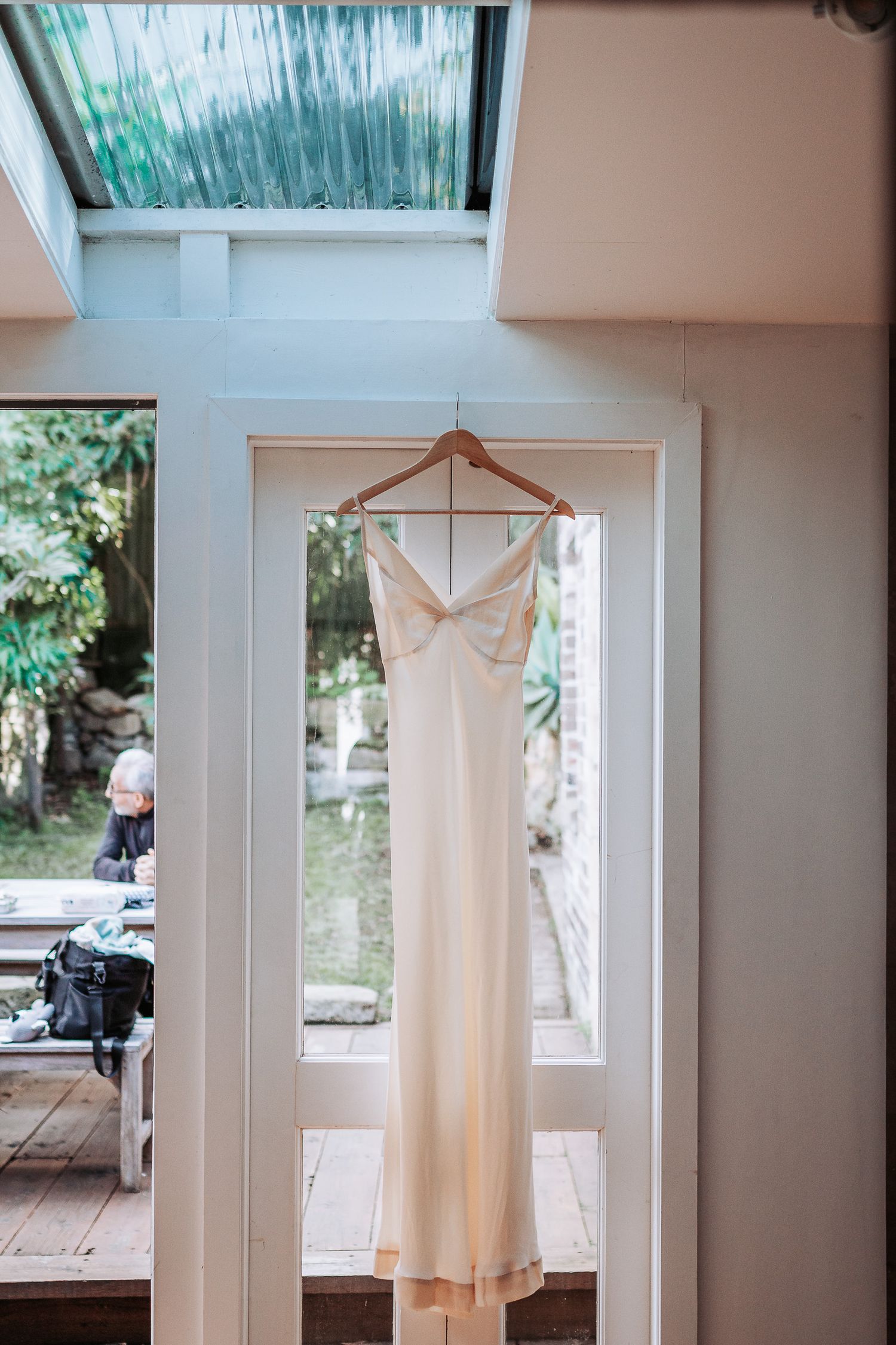 An elegant white wedding dress with spaghetti straps hangs on a wooden hanger in front of a bright window.