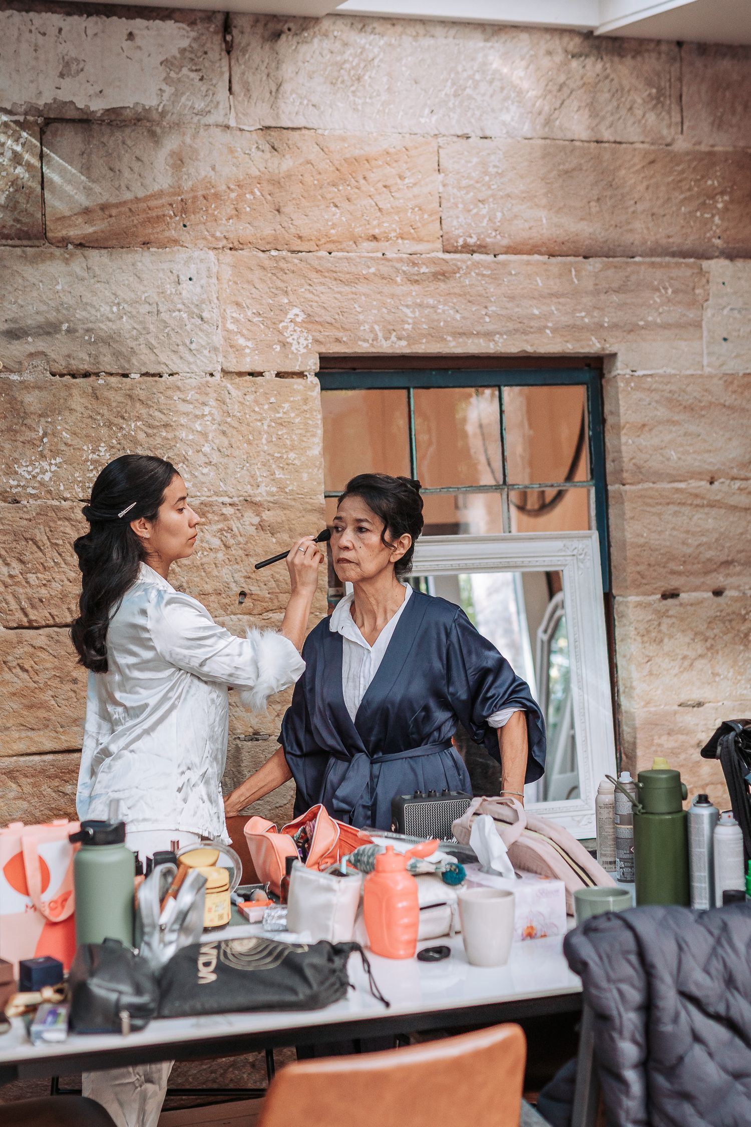 A makeup artist applies cosmetics to a client at a vanity table with various beauty products and tools.