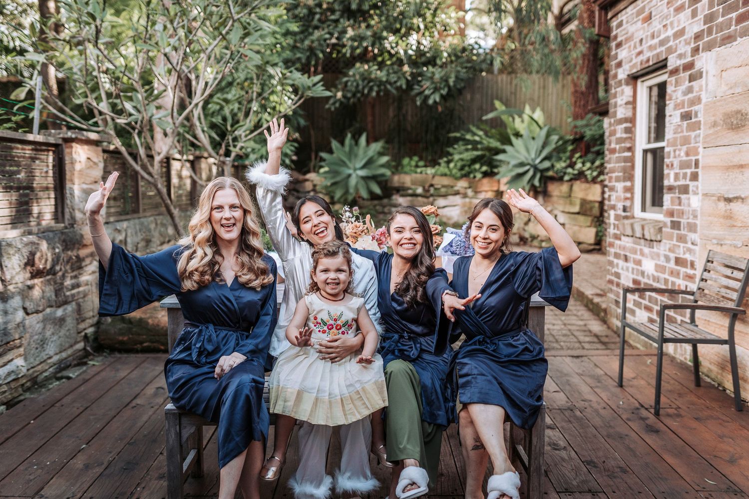 A group wearing matching navy robes sits together on outdoor wooden deck with brick wall and plants behind them.