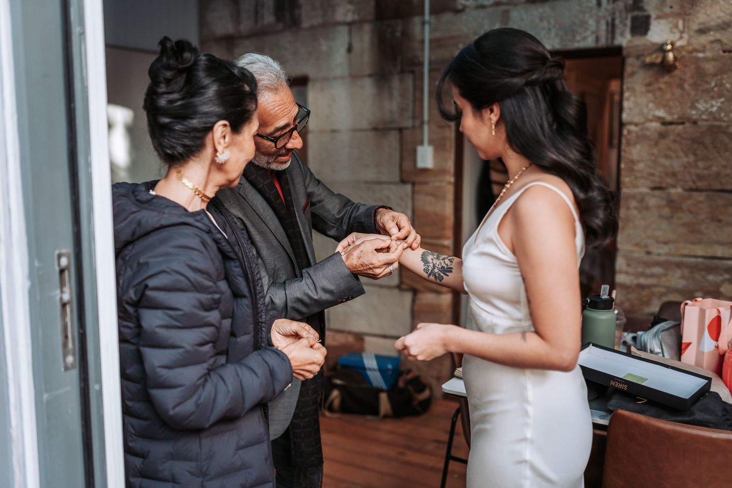 Wedding preparation scene shows someone helping with jewelry as they get ready in a cozy rustic space.