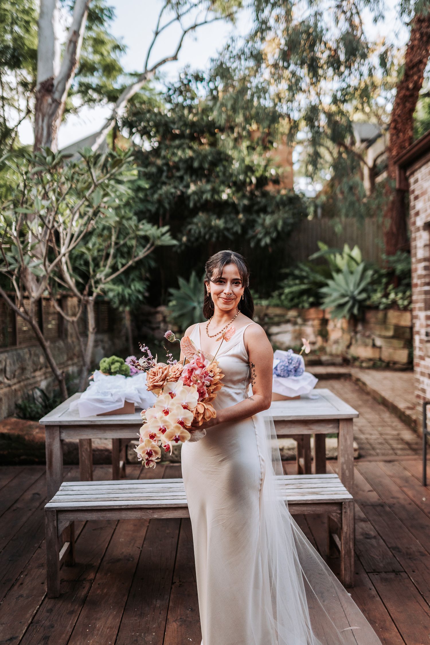 Elegant white bridal dress and bouquet of pink and peach flowers on an outdoor wooden deck surrounded by greenery.