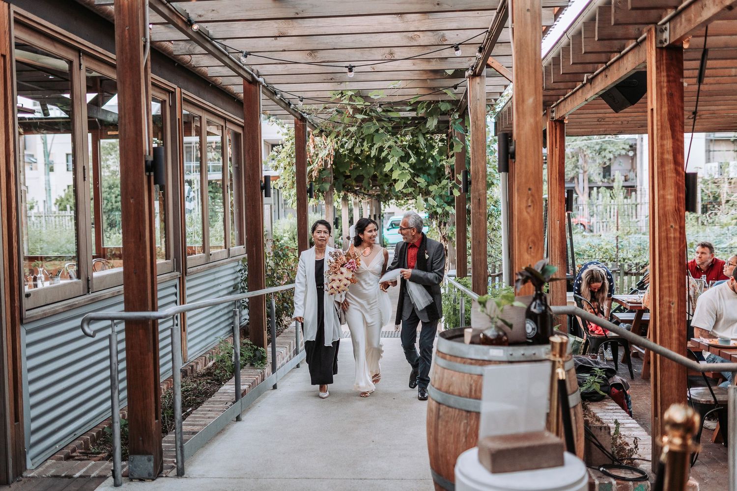 A couple and their wedding party walk down a rustic covered walkway with wooden beams and corrugated metal walls.