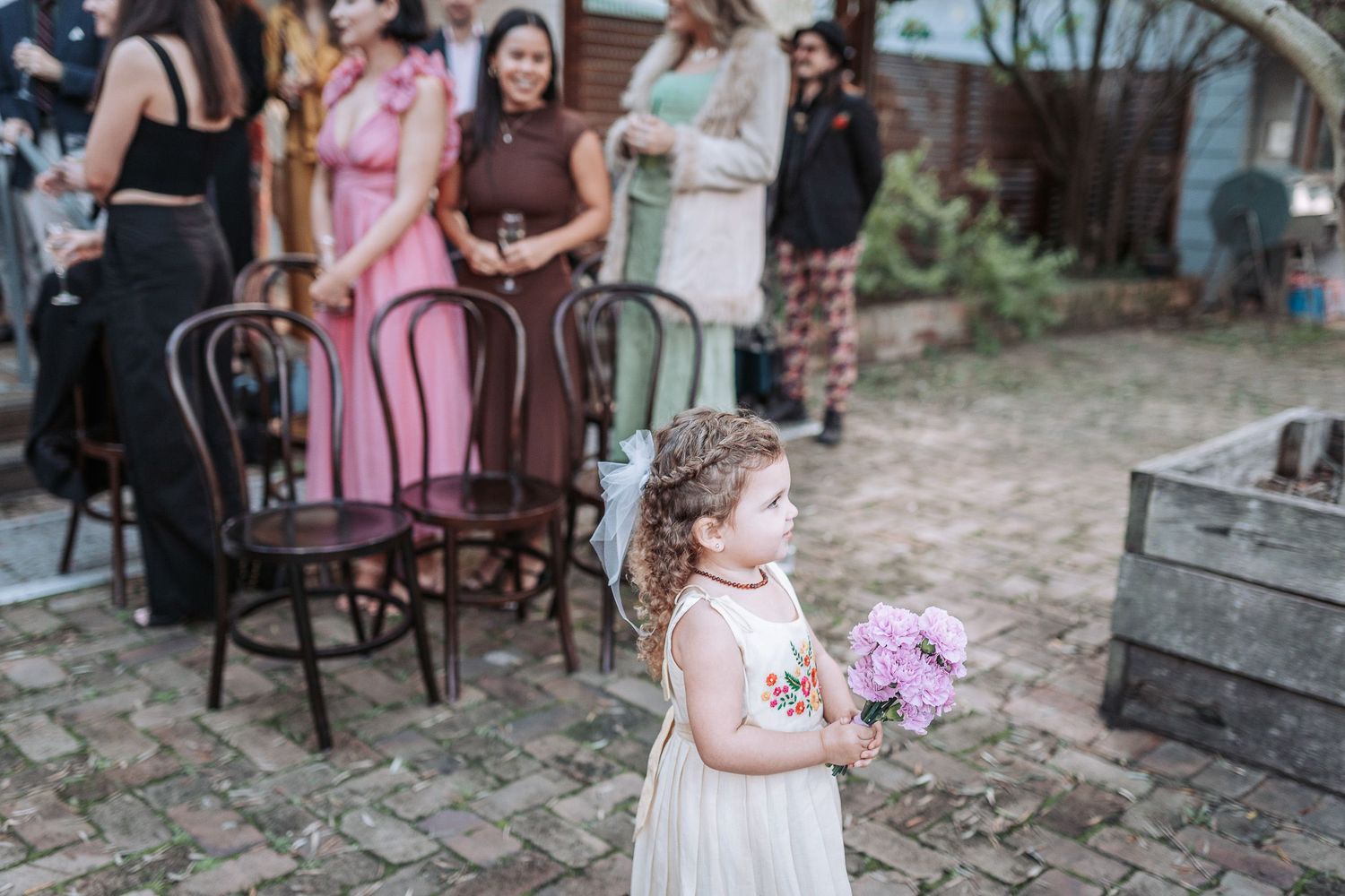 Young child in a white dress holds a bouquet of purple flowers at an outdoor gathering with bentwood chairs and guests.