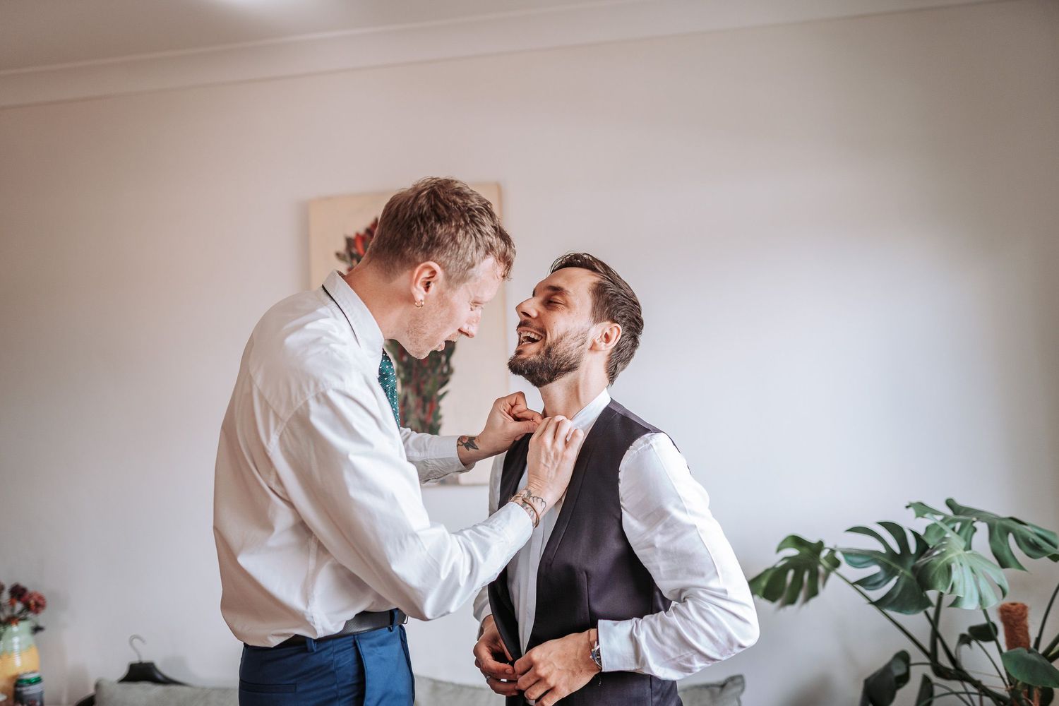 Two people help each other adjust formal attire while getting dressed in a room with houseplants.