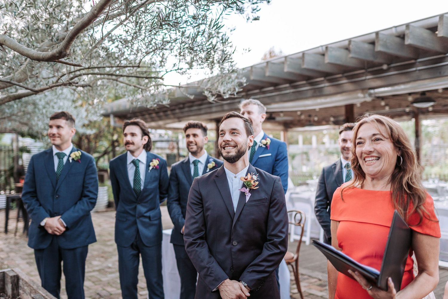 Wedding party gathers under a pergola during outdoor ceremony as officiant in coral dress delivers remarks.