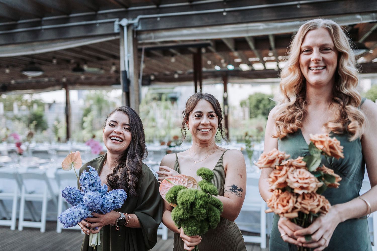 Three bridesmaids laugh joyfully while holding floral bouquets at an outdoor wedding reception venue.