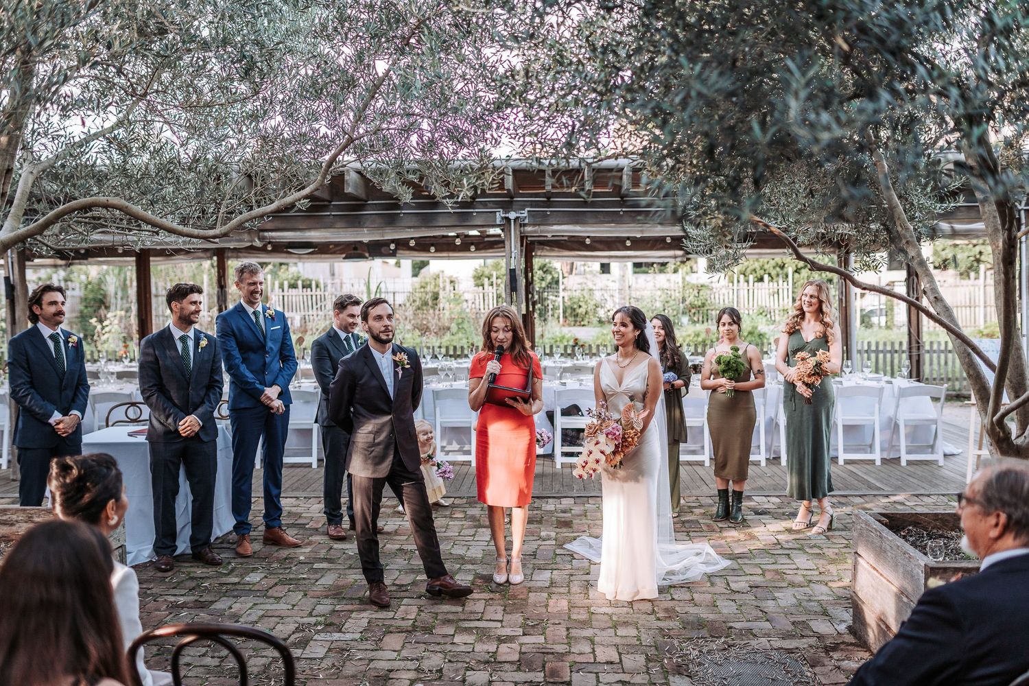 Small outdoor wedding ceremony takes place under rustic wooden arbor with guests gathered on stone patio.