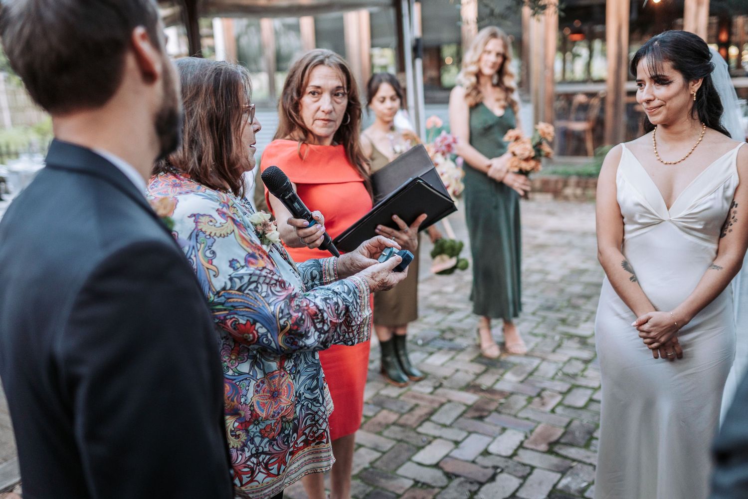 An outdoor gathering on a cobblestone street shows a ceremony officiant reading from a book while guests look on.
