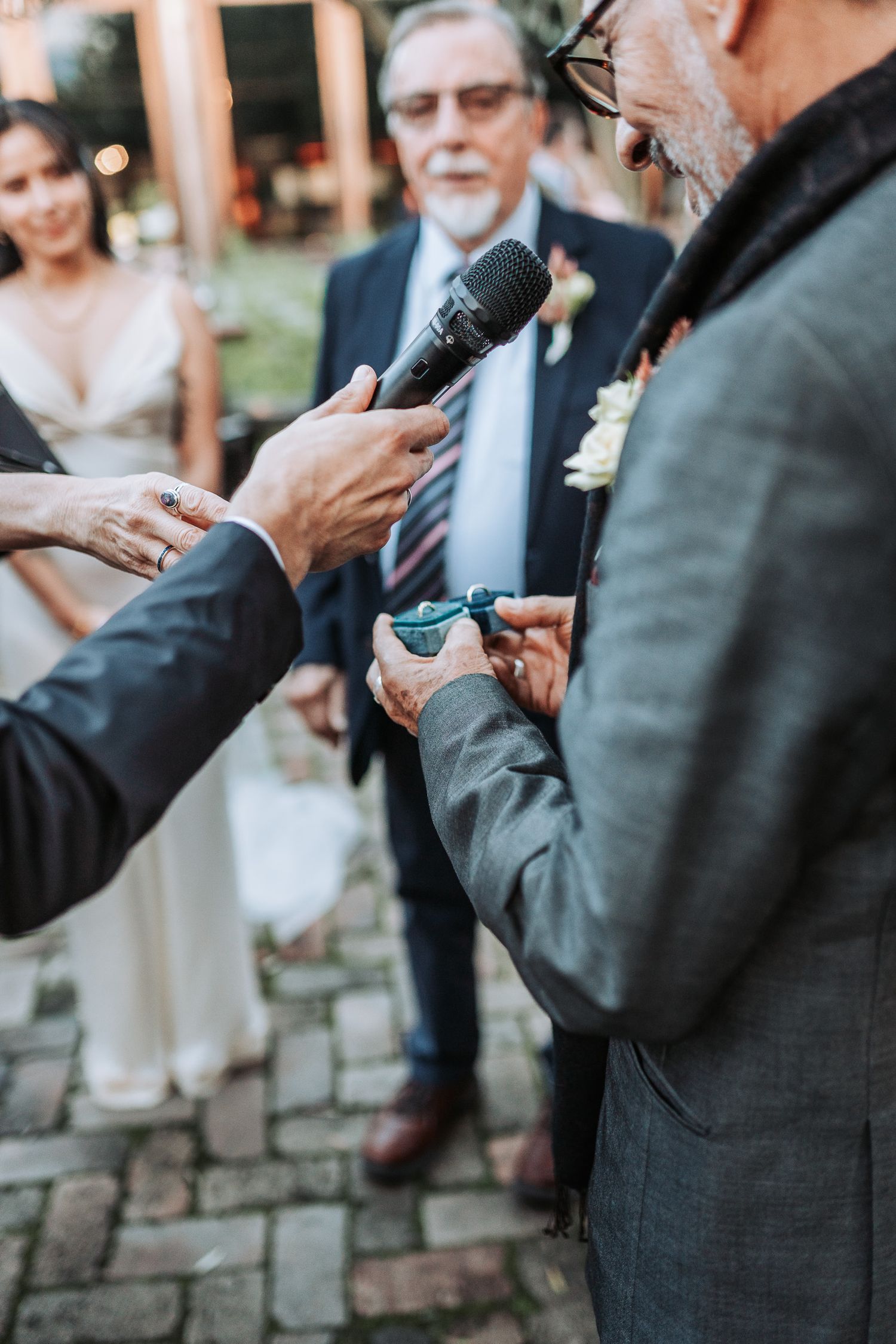 A close-up shot of people holding a microphone and glasses during an outdoor wedding ceremony on brick pavement.