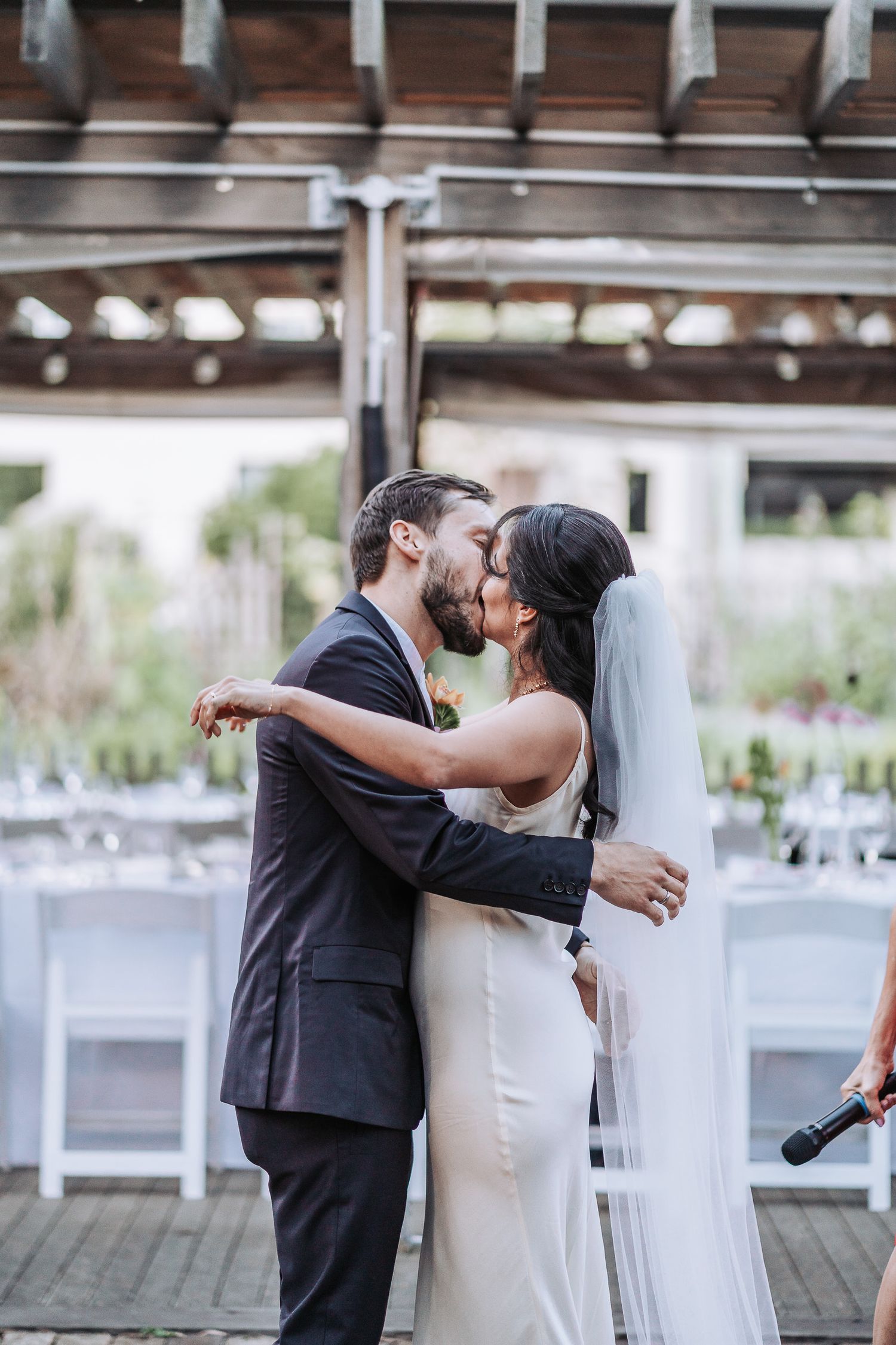 Newlyweds share their first kiss at an outdoor reception venue decorated with string lights and white chairs.