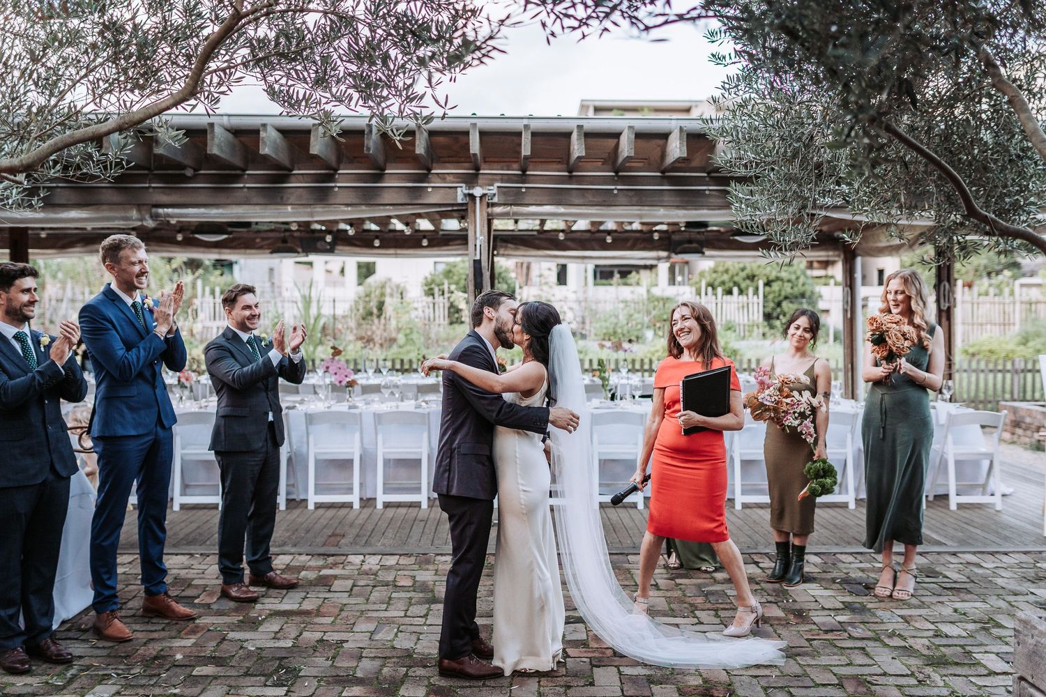 Wedding couple shares a celebratory moment under a wooden pergola as guests applaud at an outdoor reception.