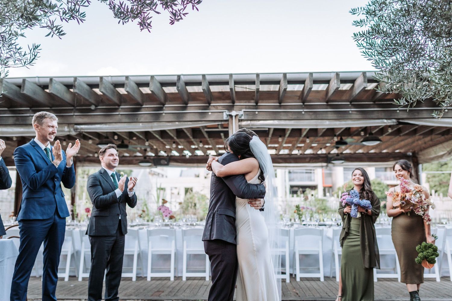 Couple shares emotional embrace during outdoor wedding ceremony while guests applaud beneath wooden pergola structure.