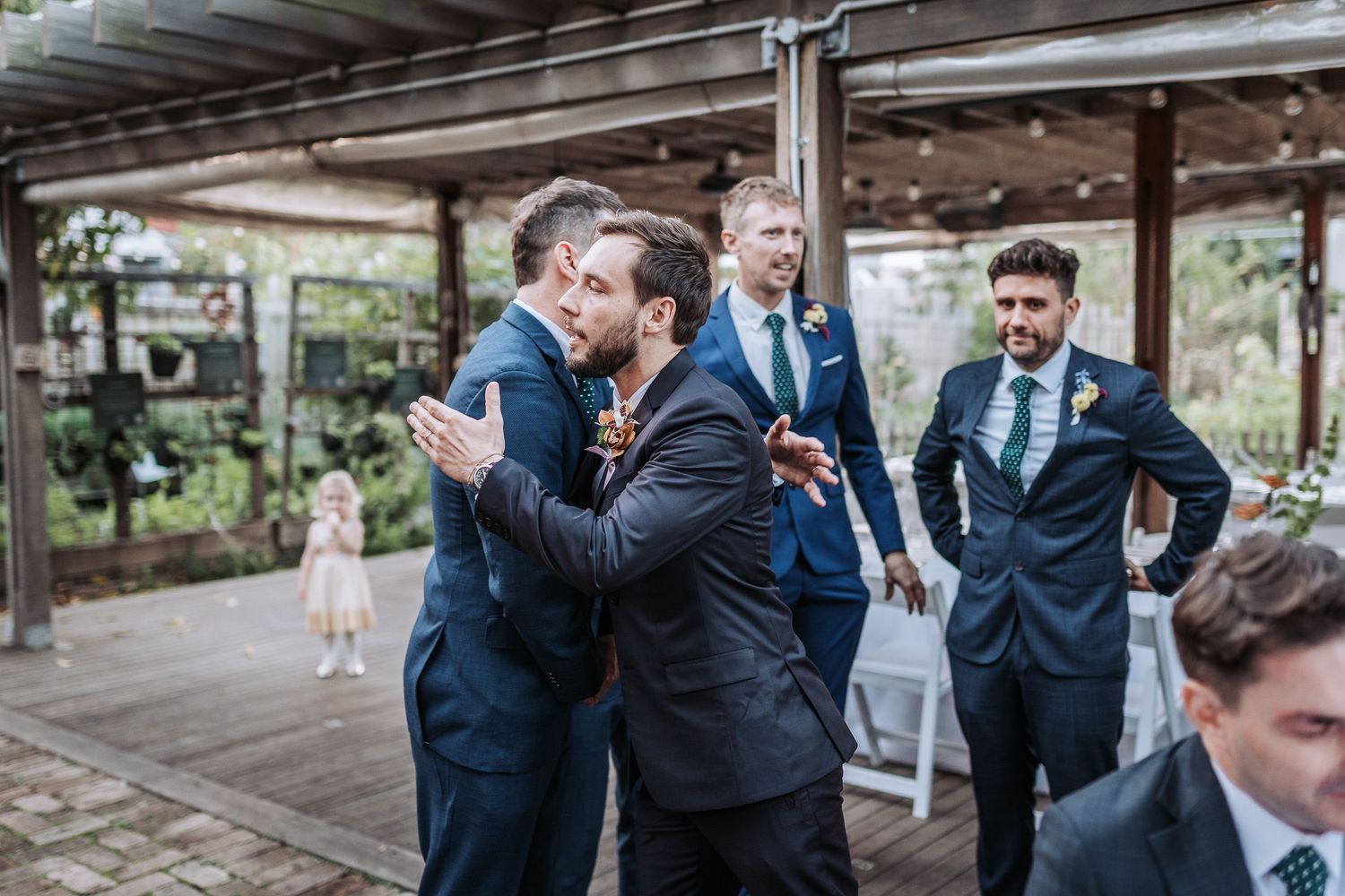 Wedding party members in navy blue suits celebrate at an outdoor venue under a covered patio area.