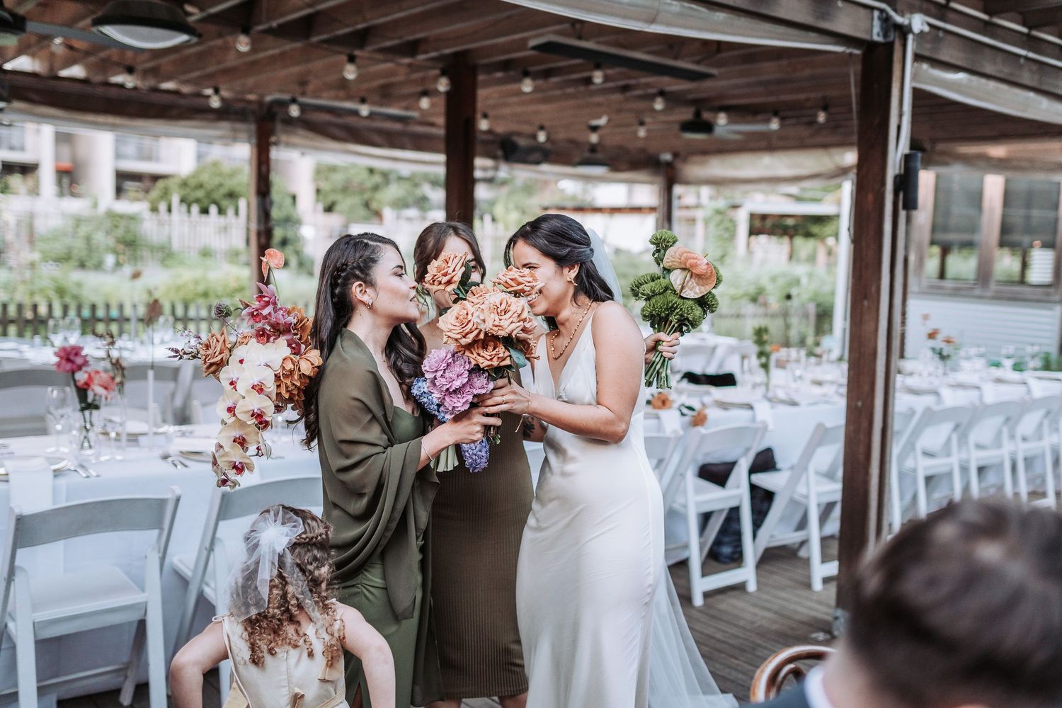 People share joyful moments at outdoor wedding reception under covered patio with floral decorations and white chairs.