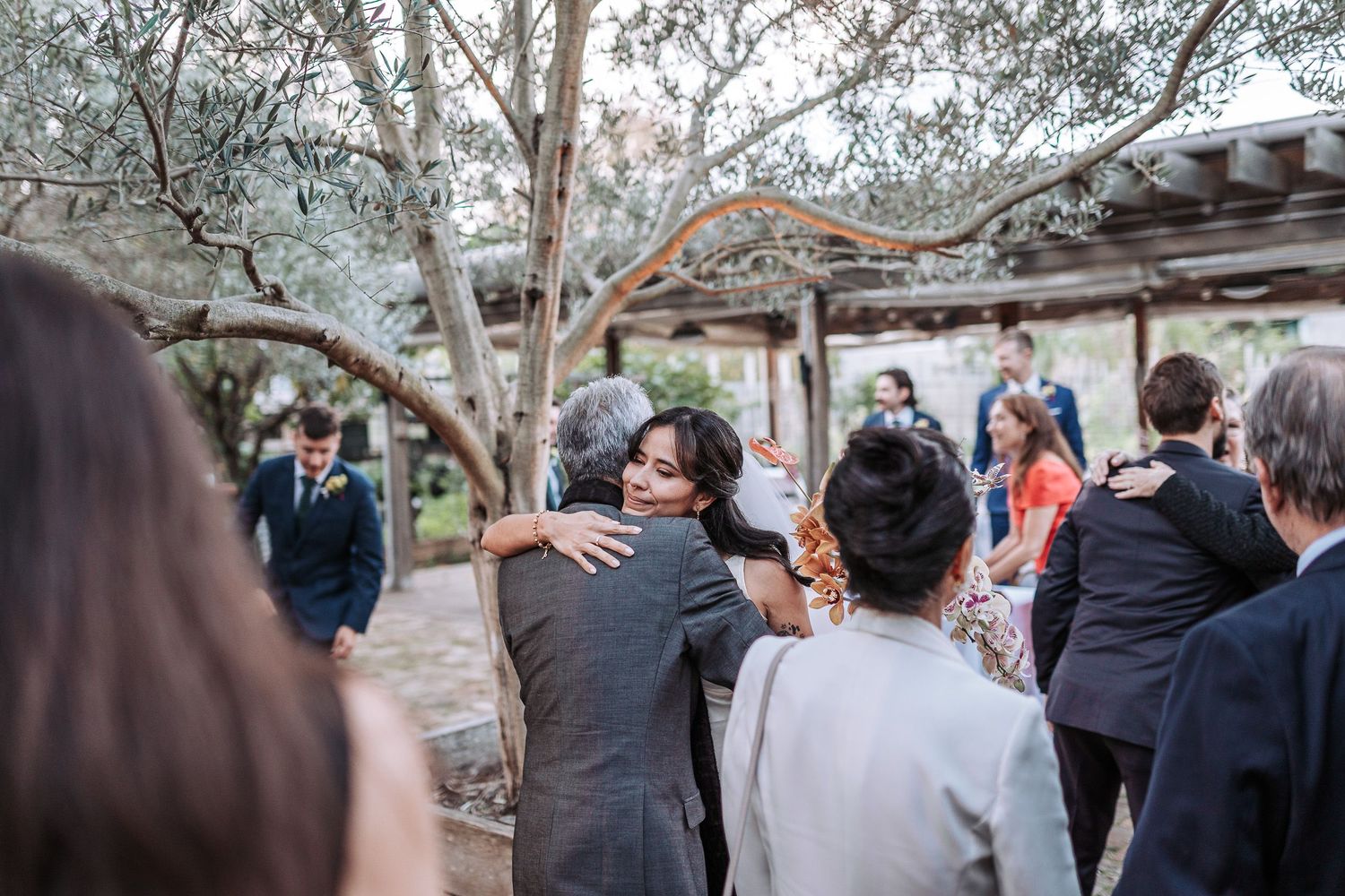 Wedding guests gather outdoors under trees during a garden ceremony while watching from a distance.