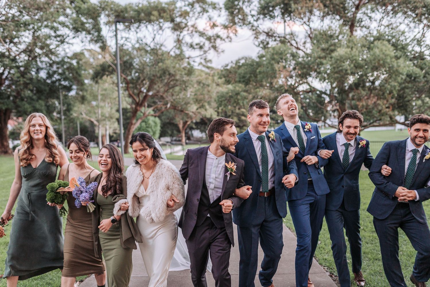 Wedding party walking together outdoors on grass with trees in background, laughing and celebrating joyfully.