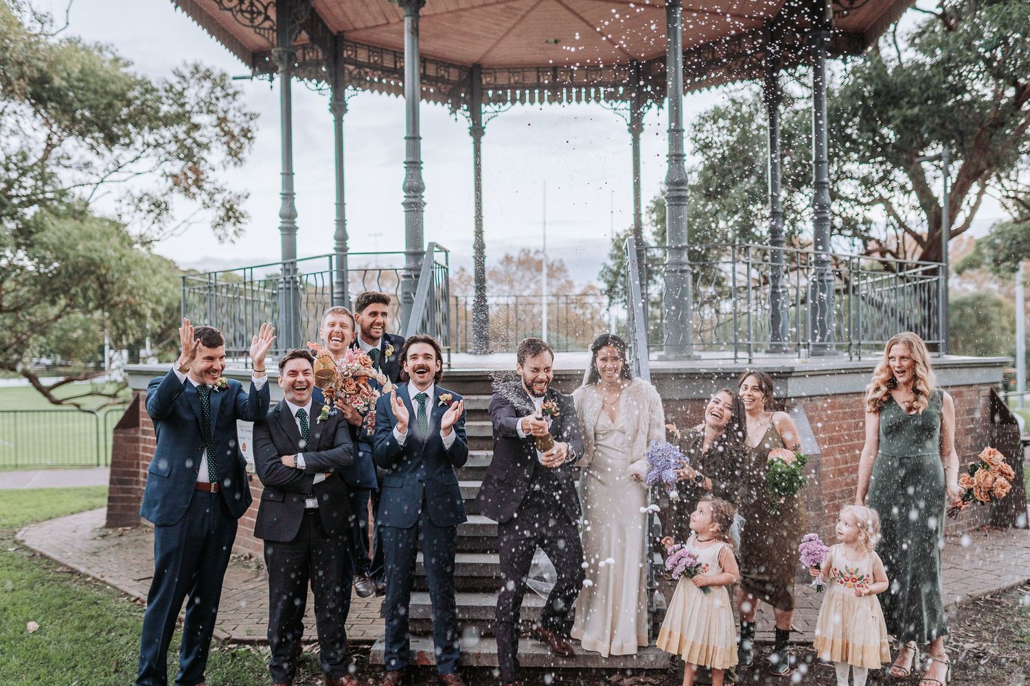 Wedding party poses together under an ornate gazebo in a park with city buildings visible in the background.