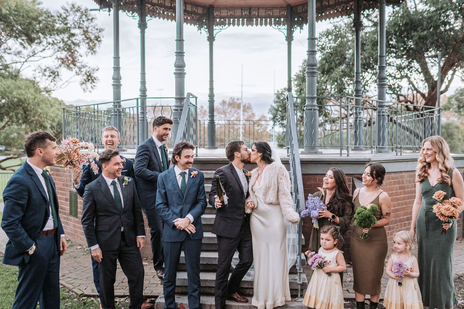 A joyful wedding party gathers under a decorative gazebo in a garden setting on a sunny afternoon.