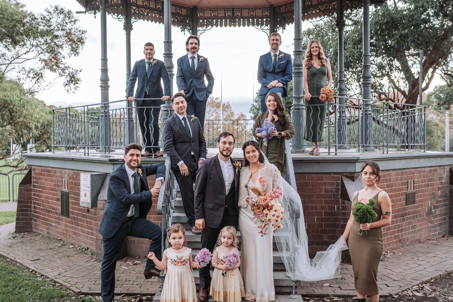 Wedding party posed together on and around a vintage brick gazebo in a park setting.
