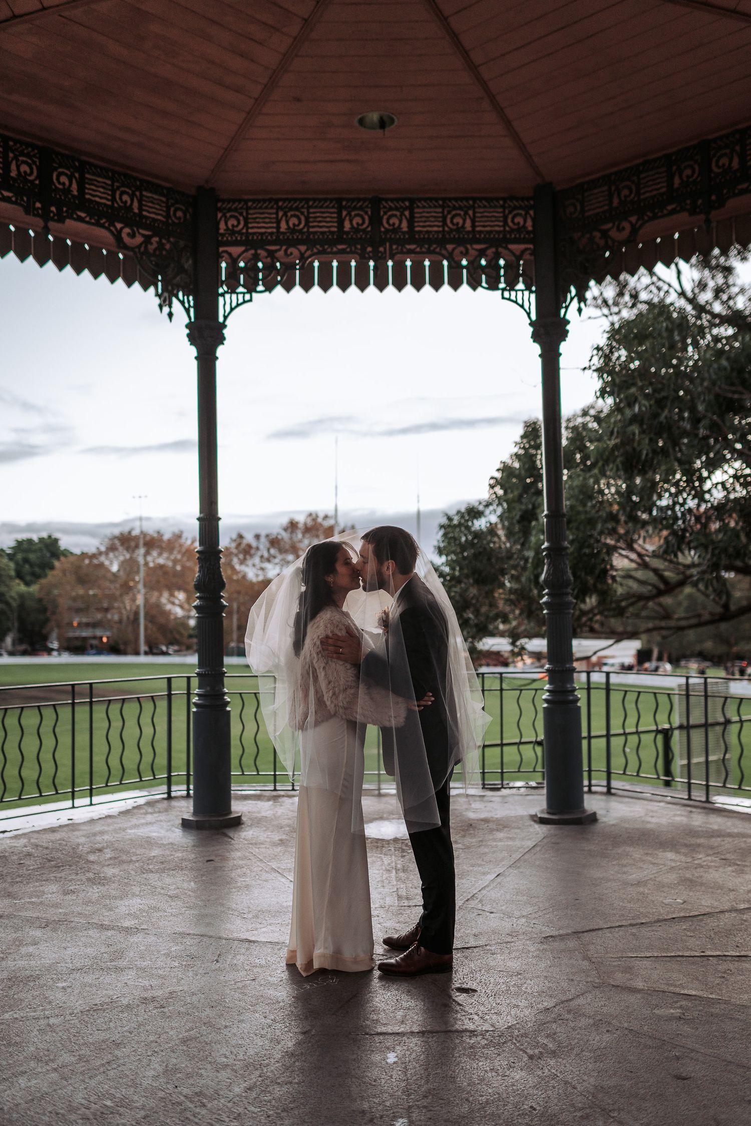 A romantic moment under an ornate Victorian gazebo in a park during autumn with overcast skies.