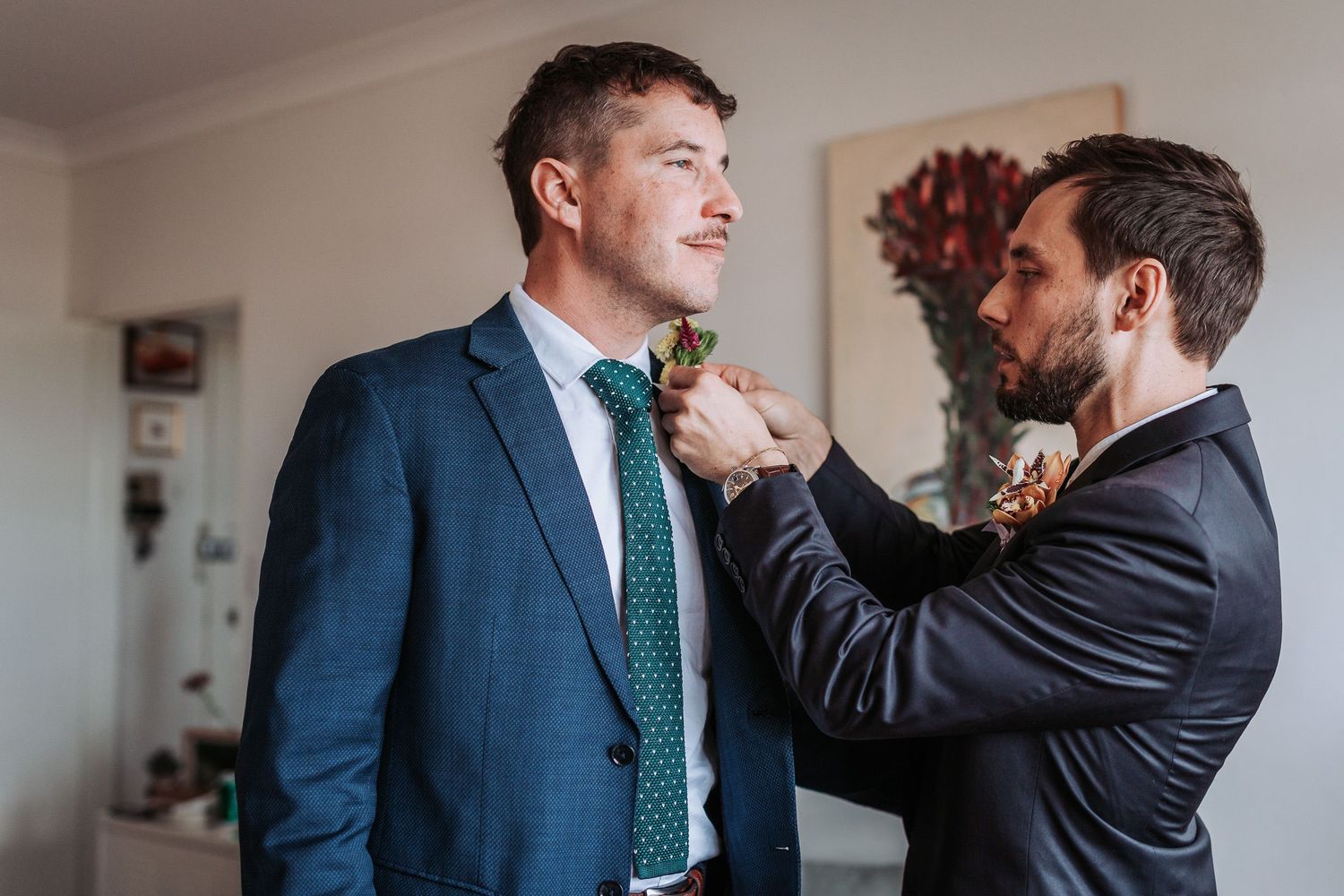 Someone helps adjust another person's tie while both wear formal suits in preparation for a special event.