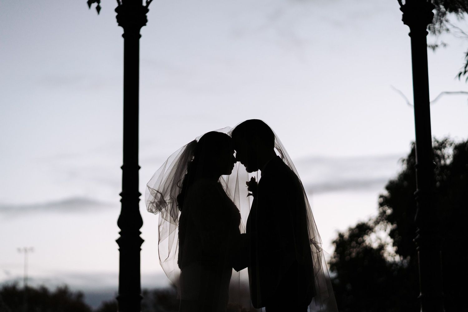 A silhouetted couple in wedding attire sharing an intimate moment beneath a lamp post at dusk.