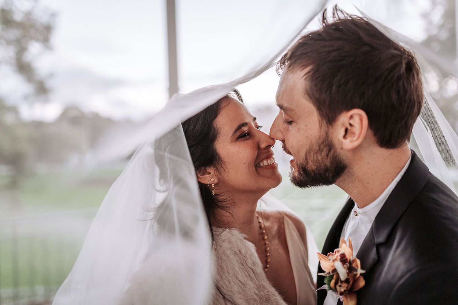 A joyful wedding couple sharing an intimate moment together under a veil during their outdoor ceremony.