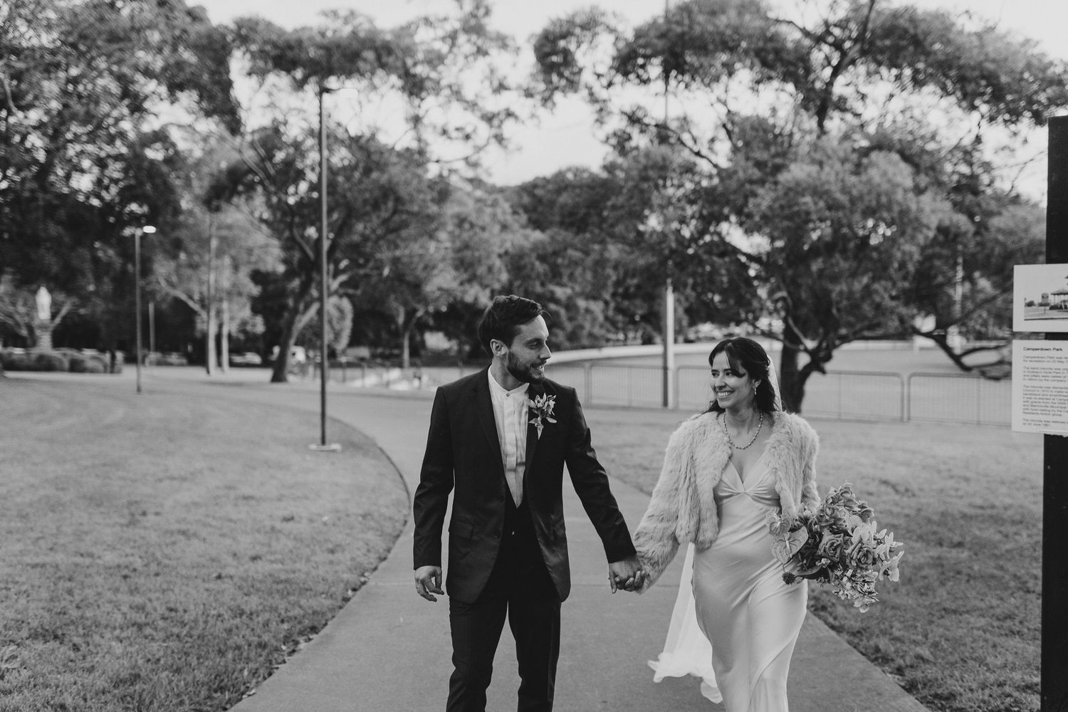 A couple in wedding attire walks hand in hand through a park with trees in black and white photography.