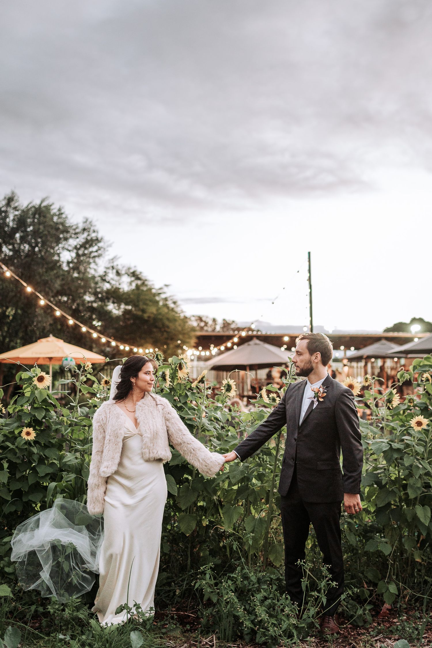 A couple holds hands in a sunflower field under string lights during a romantic outdoor wedding celebration.