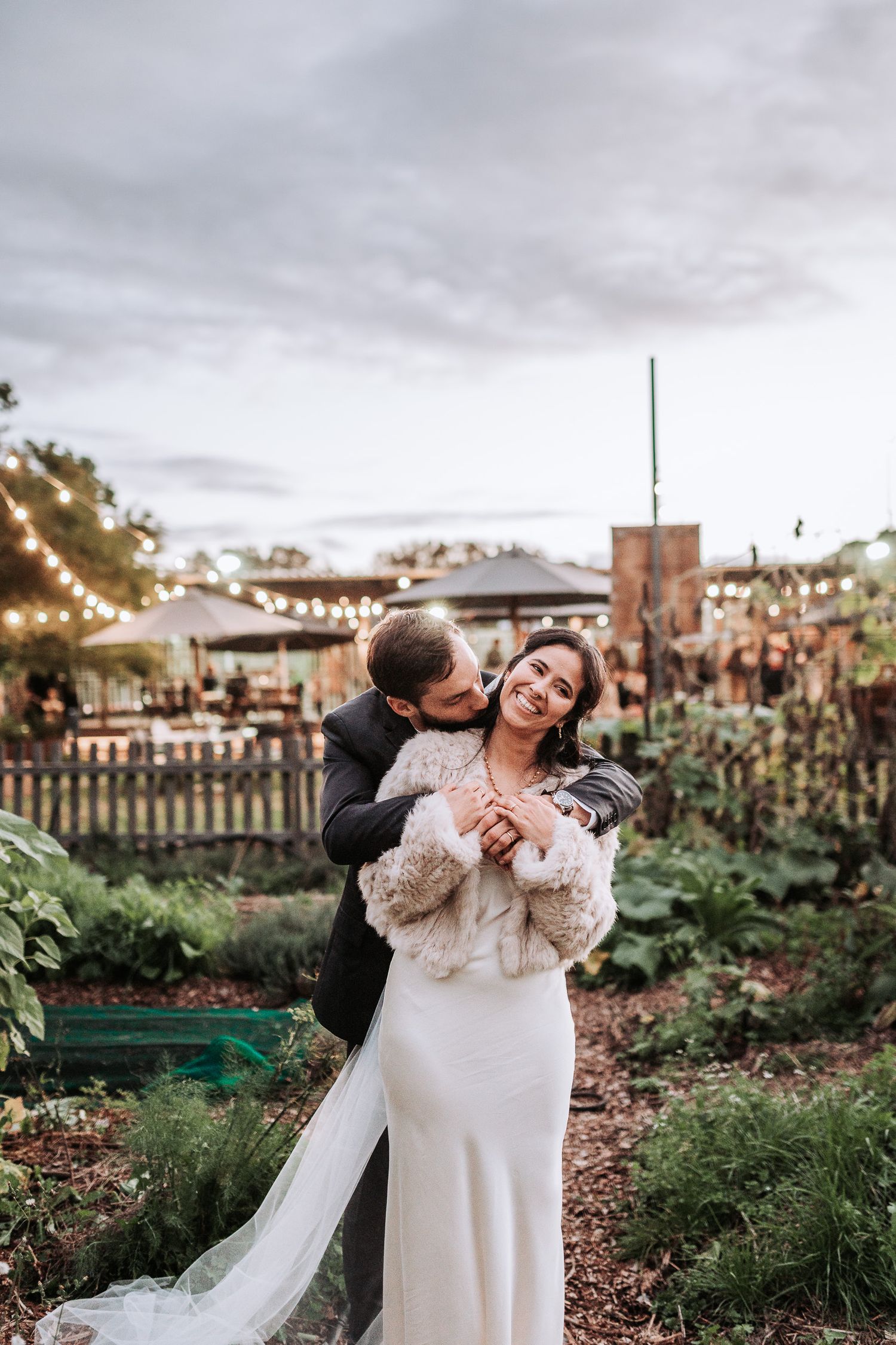 A couple shares a joyful moment in a garden setting with string lights and greenery during their wedding celebration.