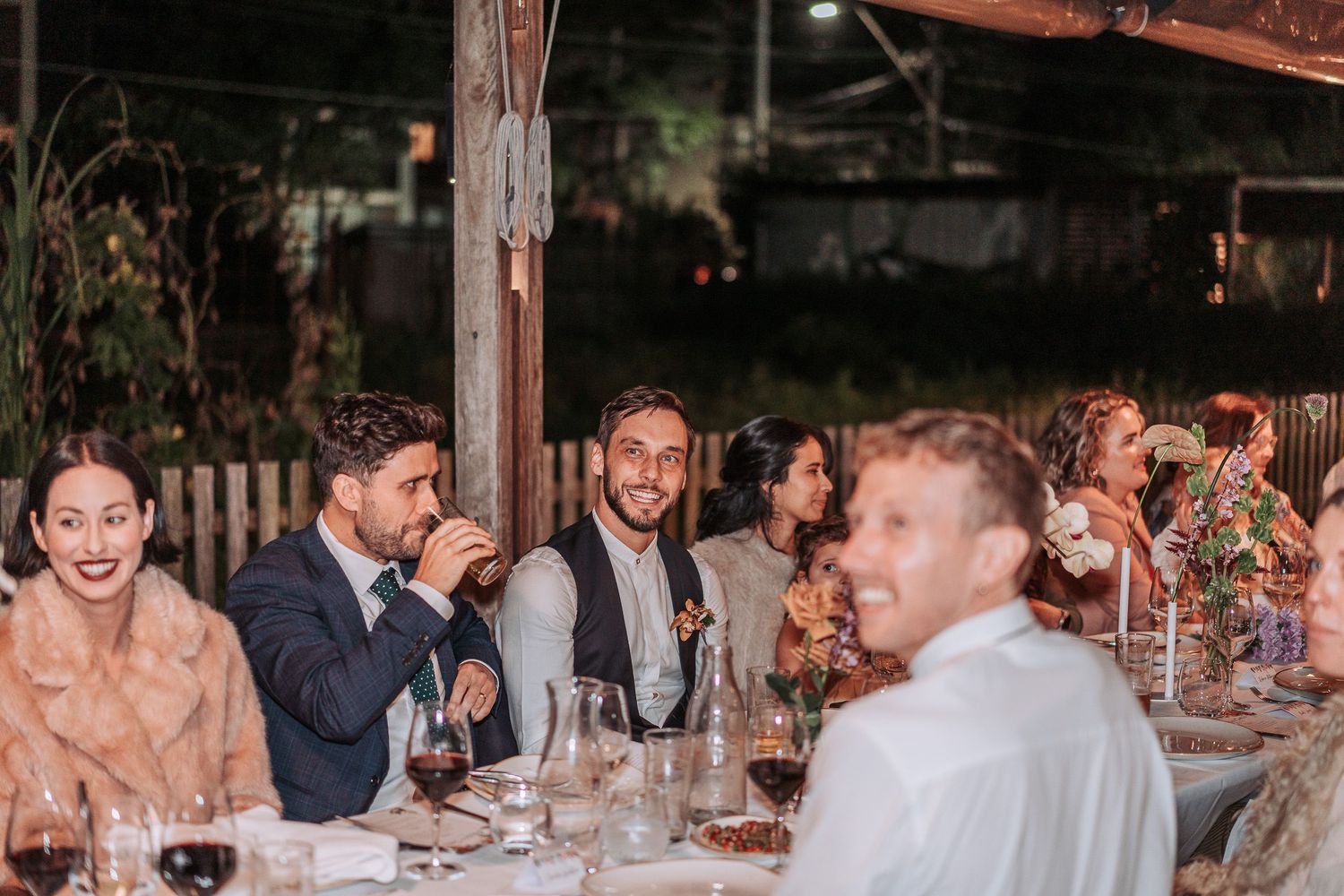 A group of guests enjoying dinner and conversation at a rustic outdoor reception with string lights overhead.