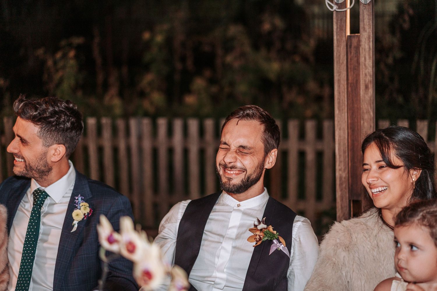 Three wedding guests share laughter during an outdoor celebration with a wooden fence in the background.