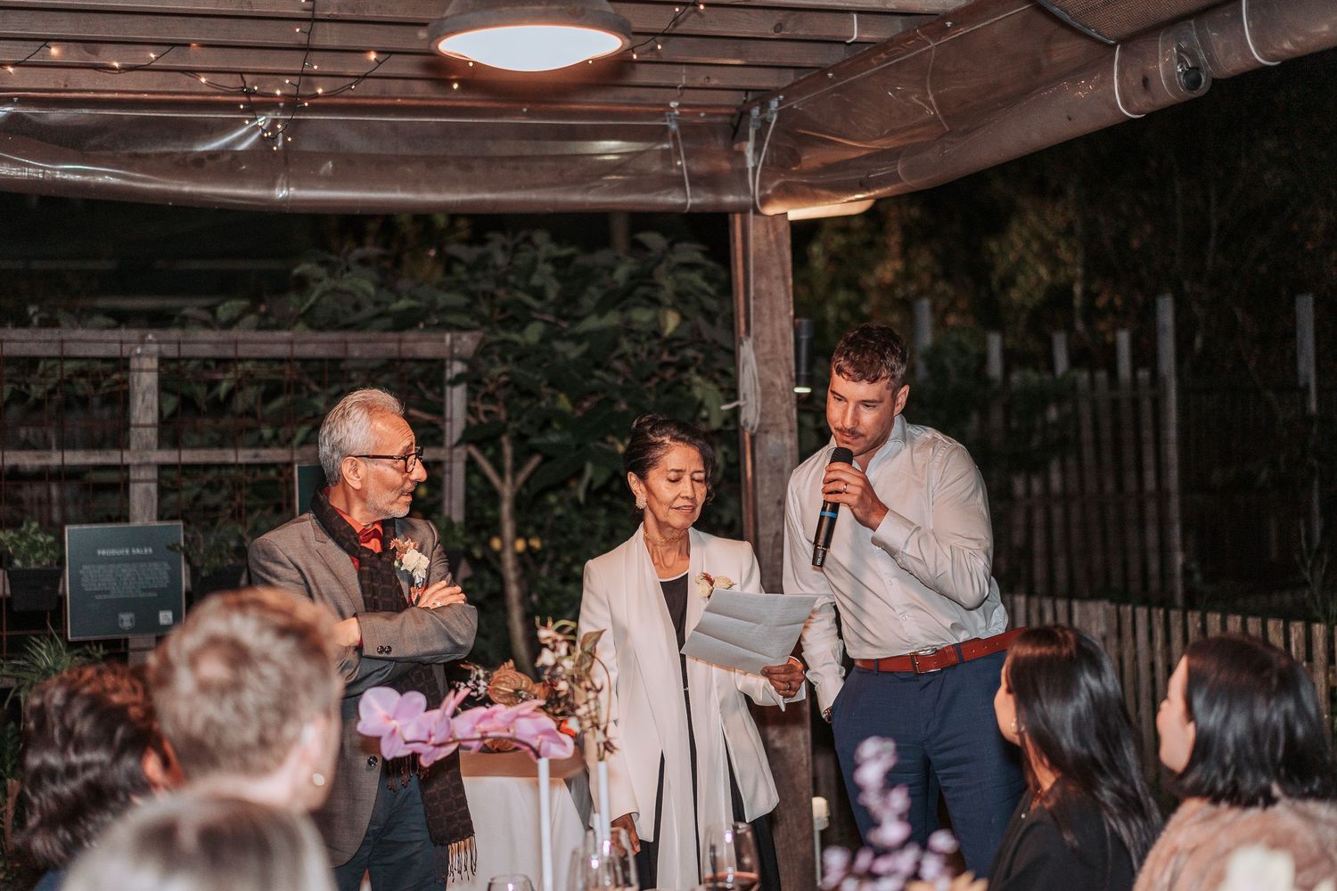 Evening outdoor wedding reception with guests gathered under wooden beams and string lights during speeches.