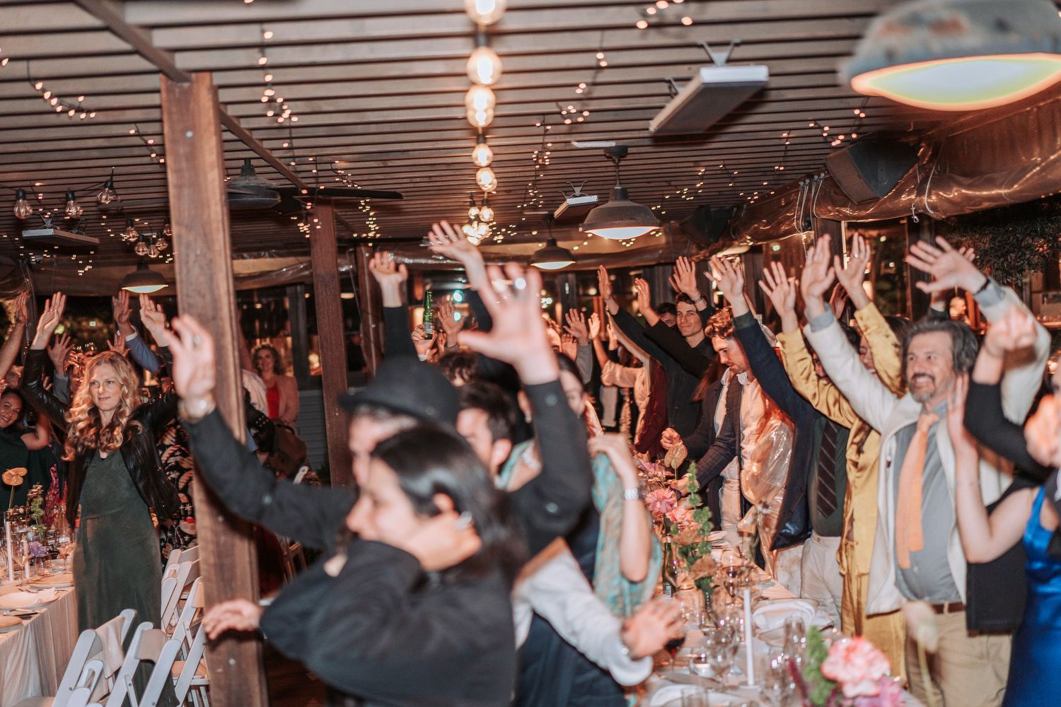 Crowd of wedding guests with raised hands celebrating energetically at an indoor reception venue with overhead lighting.