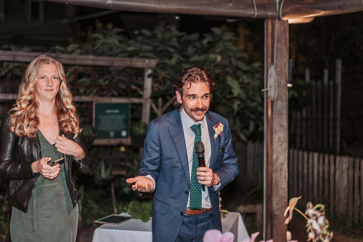 Two wedding attendees stand at a rustic outdoor venue as they make heartfelt toasts with glasses in hand.