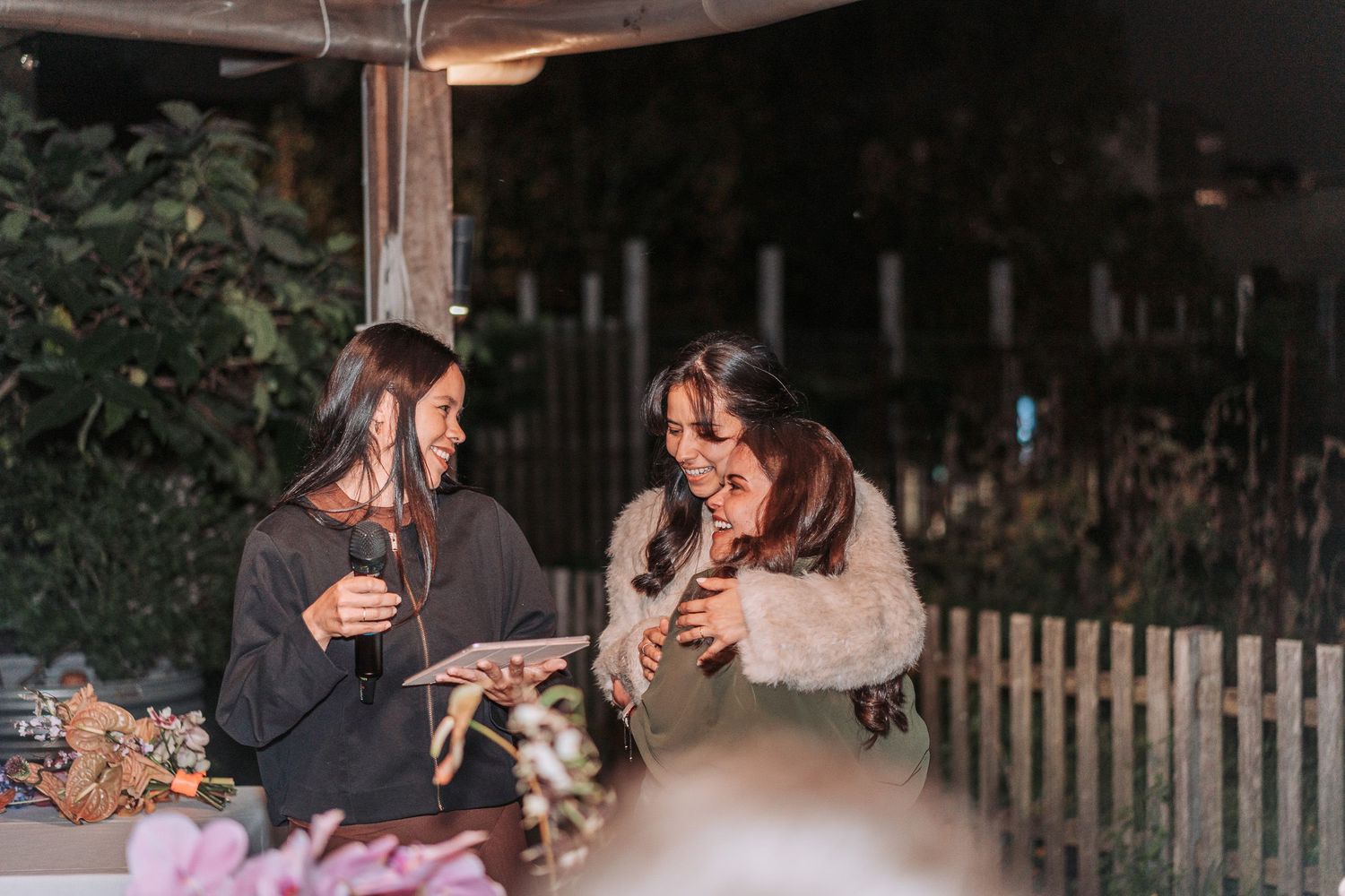 Two friends enjoying drinks and conversation at a cozy outdoor gathering with string lights and a rustic fence.