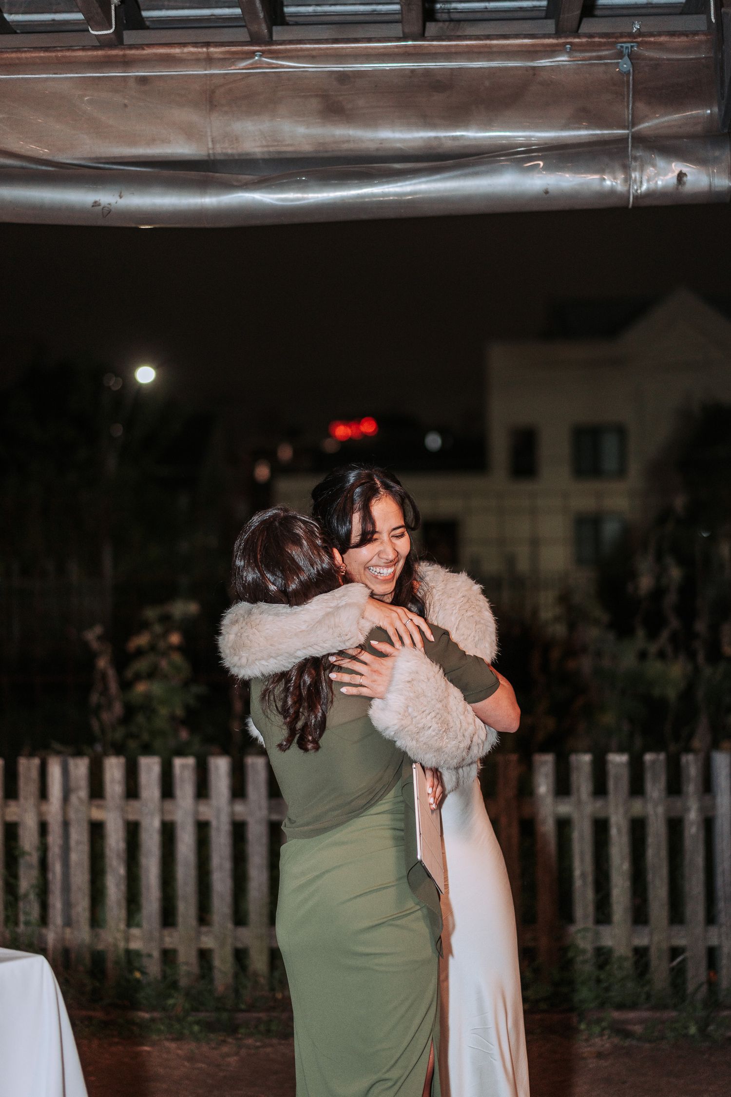 Two people share a warm embrace at an outdoor evening reception with a white picket fence in the background.