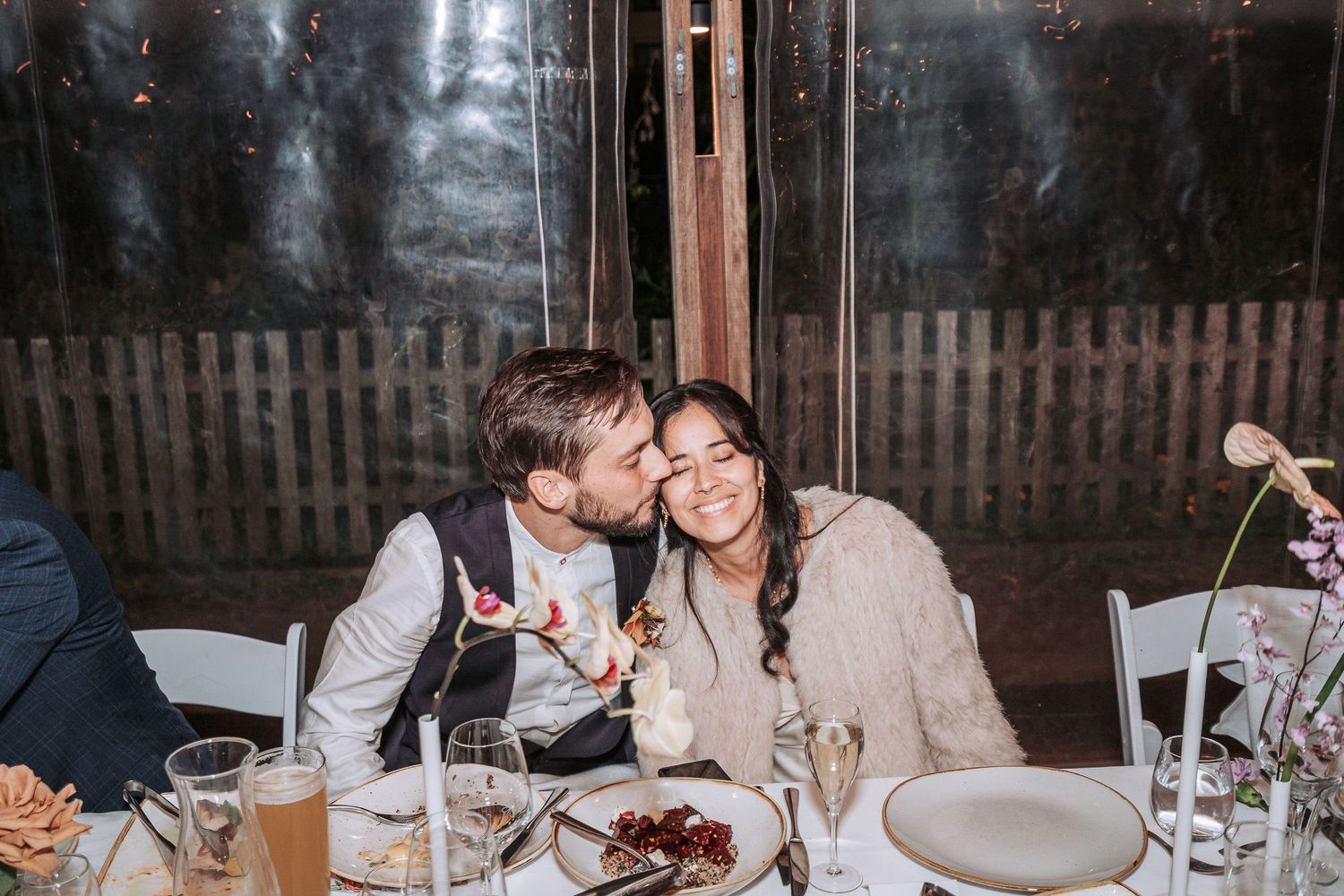Two people in formal attire share an intimate moment at a dinner table with wine glasses and plates visible.