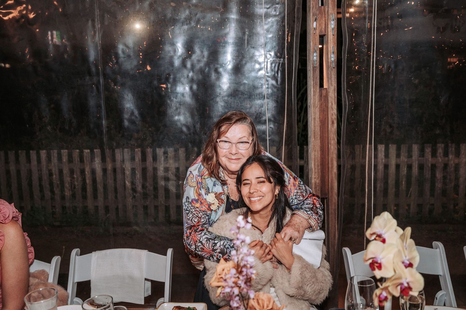 Two people sitting at white table at outdoor evening event with decorative lighting and floral centerpieces.