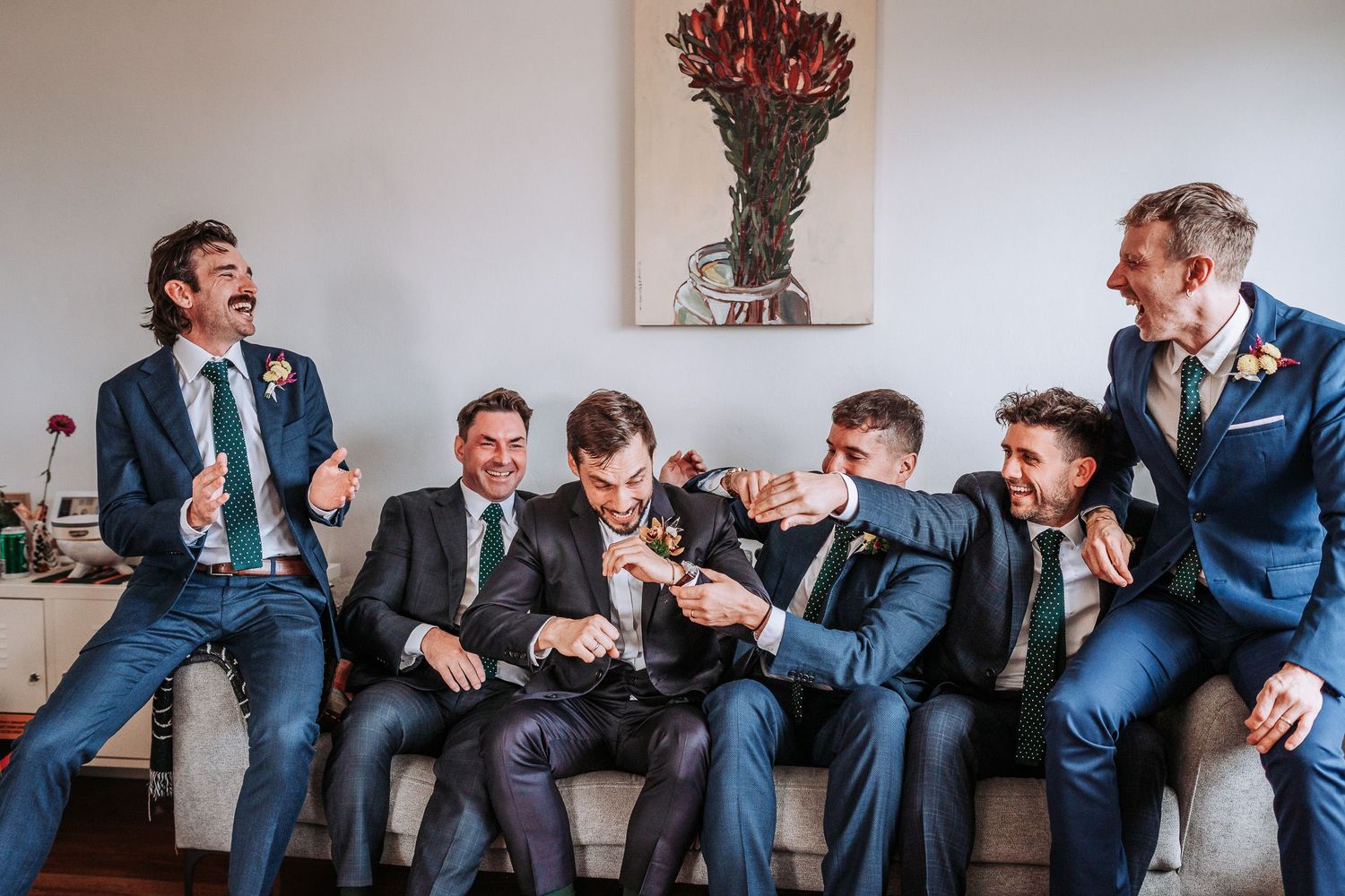 A group of groomsmen in navy suits laugh together while sitting on a couch under floral wall art.