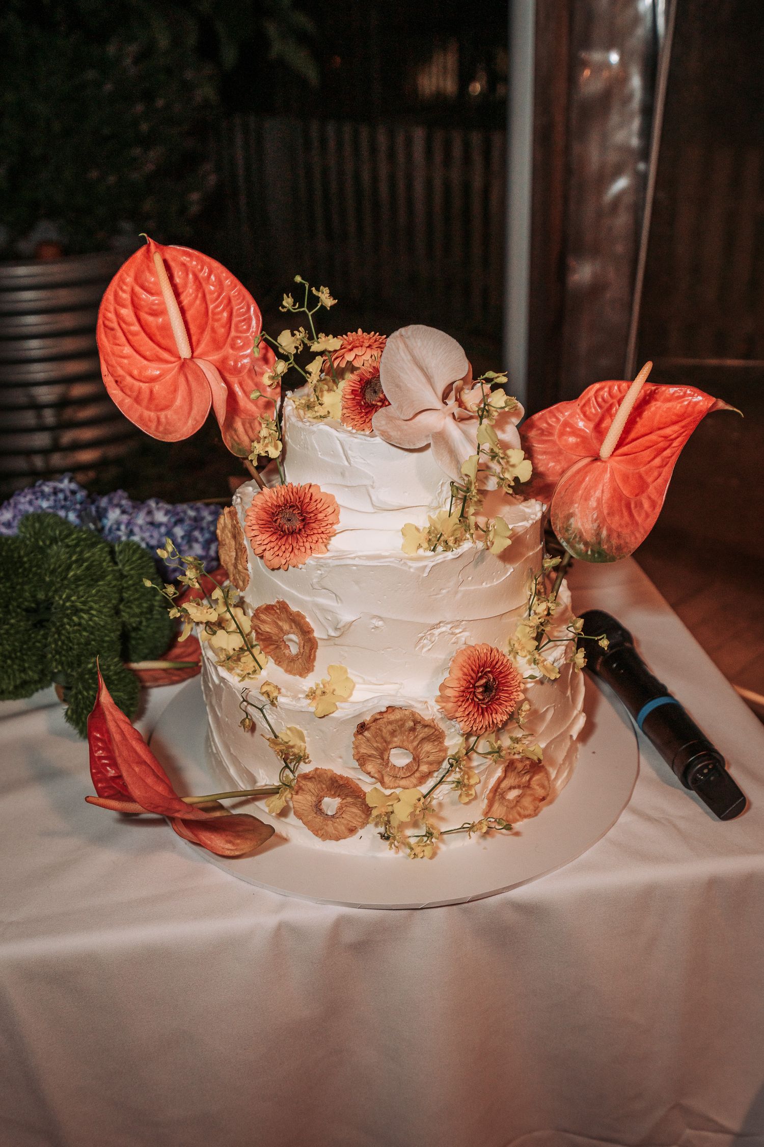 Elegant white wedding cake decorated with coral anthurium flowers, pink gerbera daisies, and golden accents on a table.
