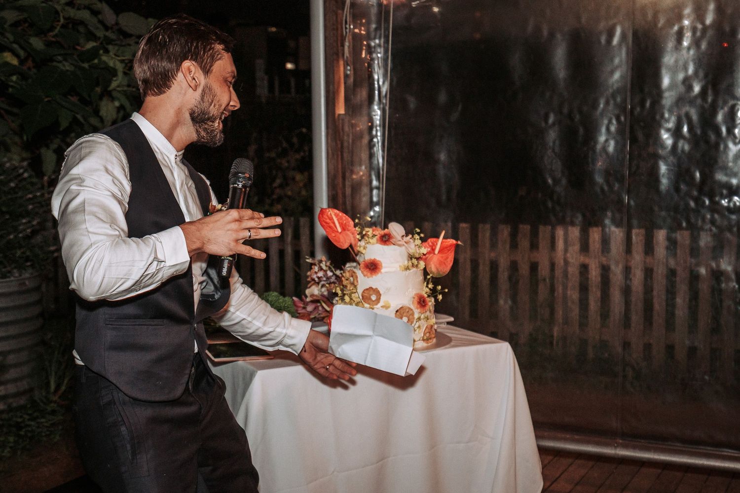 A person in a waistcoat and white shirt cuts an elegant white and floral wedding cake at a dimly lit reception.